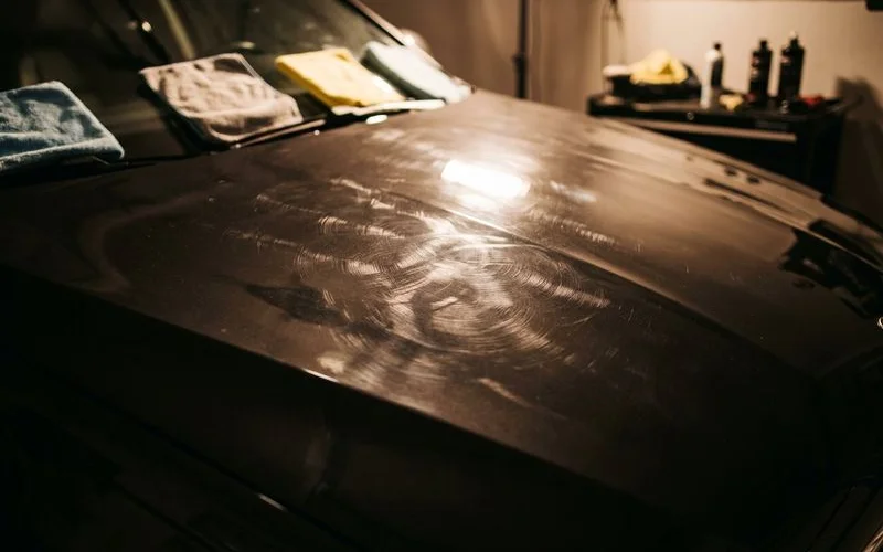 Swirl marks clearly visible on dark vehicle hood panel illuminated by direct sunlight at specific angle