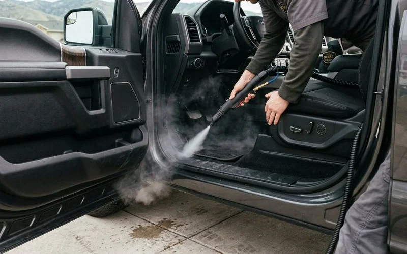 Professional interior detailing in progress with steam cleaner being used on vehicle carpet and floor mats